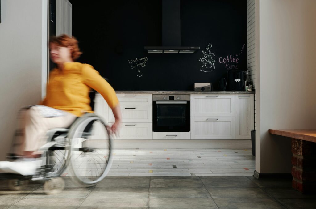 A ginger woman in a wheelchair wearing a yellow sweater is moving around her kitchen space